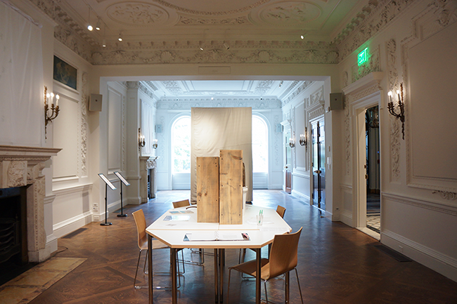 A white ballroom with elaborate plaster moulding filled with a large canvas shrine and dirty research library.